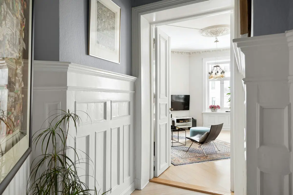 A white panelled hallway with wainscoting and a framed picture, opening through double doors to a bright living room with an armchair and a ceiling rose