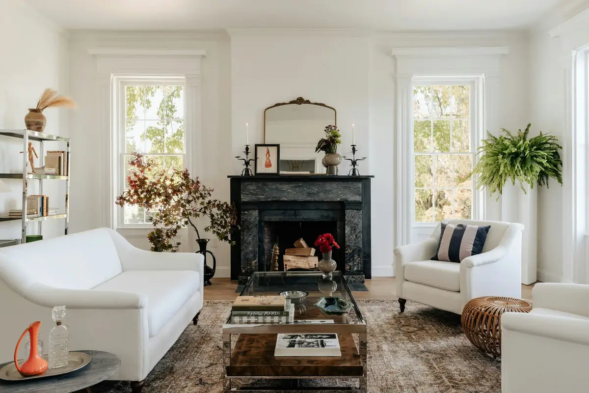 Bright, airy living room with two tall white sash windows, panelled architraves, a dark marble fireplace with a gilt mirror above it, a white sofa and armchairs on a worn rug