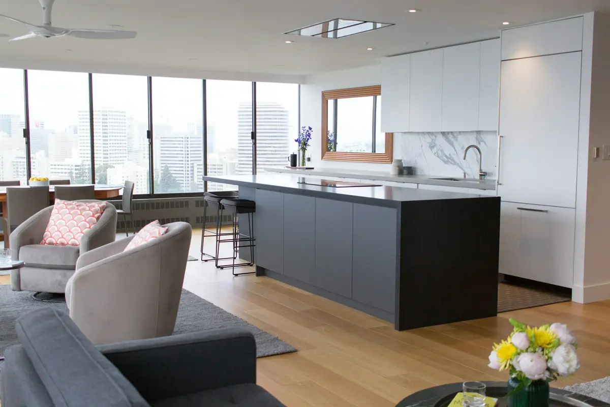 Open-plan apartment with a dark grey island kitchen, white cabinetry, two soft armchairs and floor-to-ceiling windows overlooking a city skyline