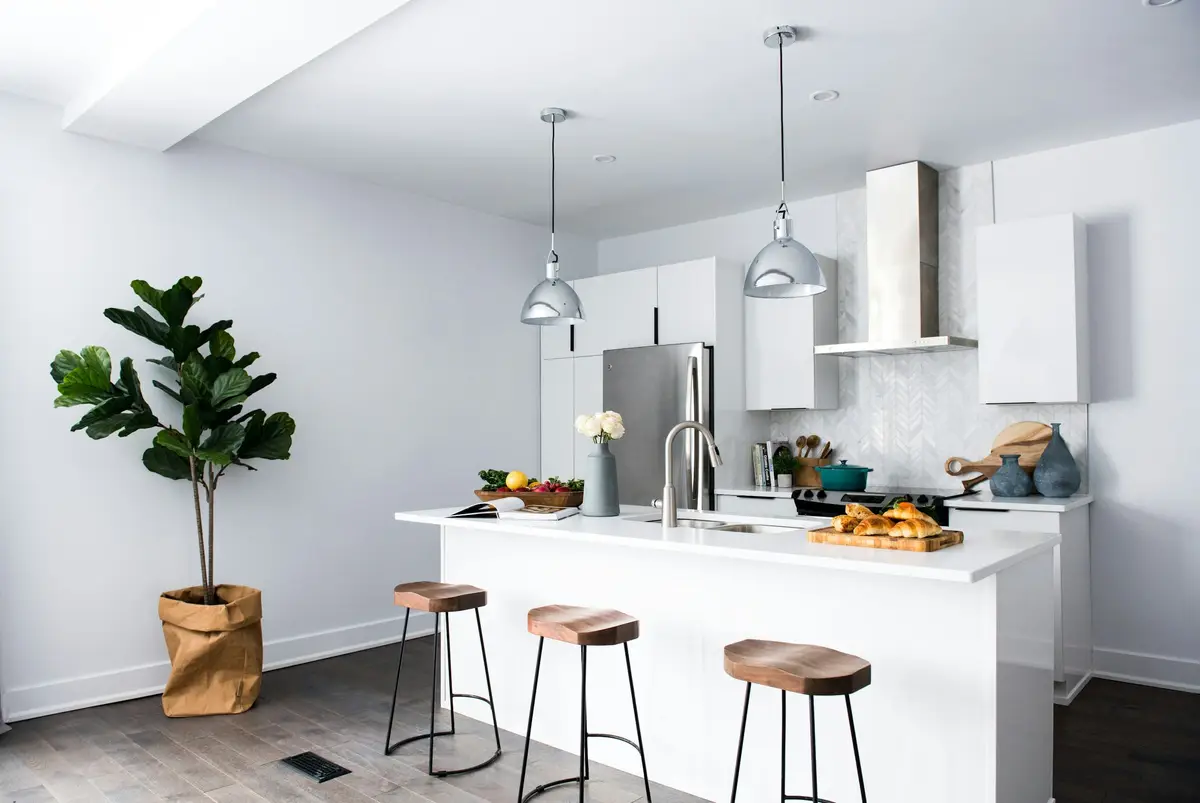 Bright modern kitchen with a white breakfast bar, three wooden-seat bar stools, steel pendant lights, an extractor hood and a fiddle-leaf fig in a paper bag planter