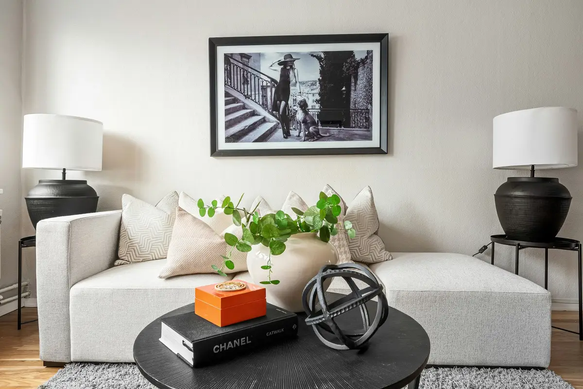 Compact living room with a pale sofa, a round black coffee table holding books and a plant, two black table lamps and a framed black-and-white print