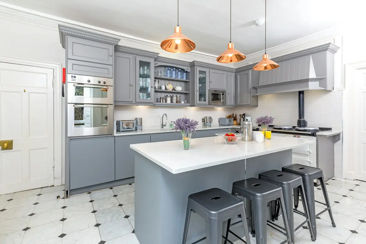Country-style kitchen with grey painted cabinets, a central island with metal bar stools, copper pendant lights, a double oven and a black-and-white tiled floor