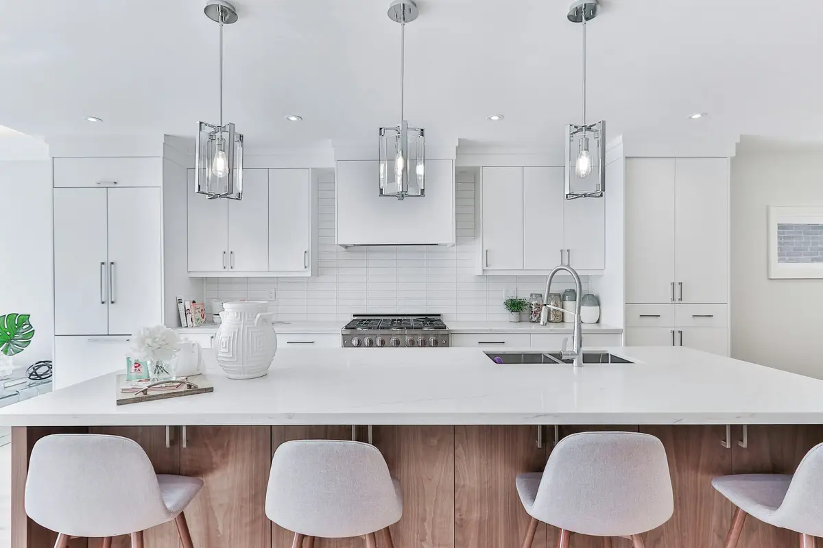 White kitchen with a wood-panelled island and four white stools, glass pendant lights, a range cooker and a white subway-tile splashback