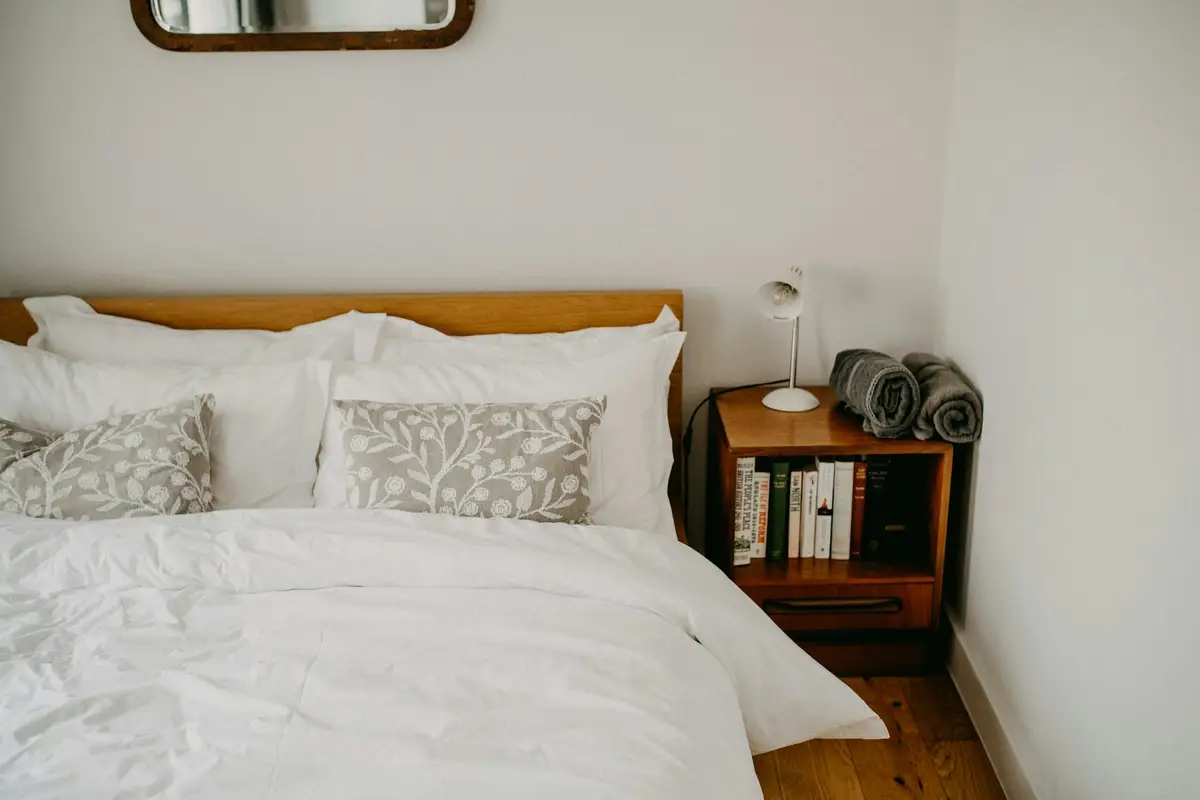 A bed with white linen and patterned cushions beside a wooden bedside cabinet holding books and a small lamp, with a mirror above
