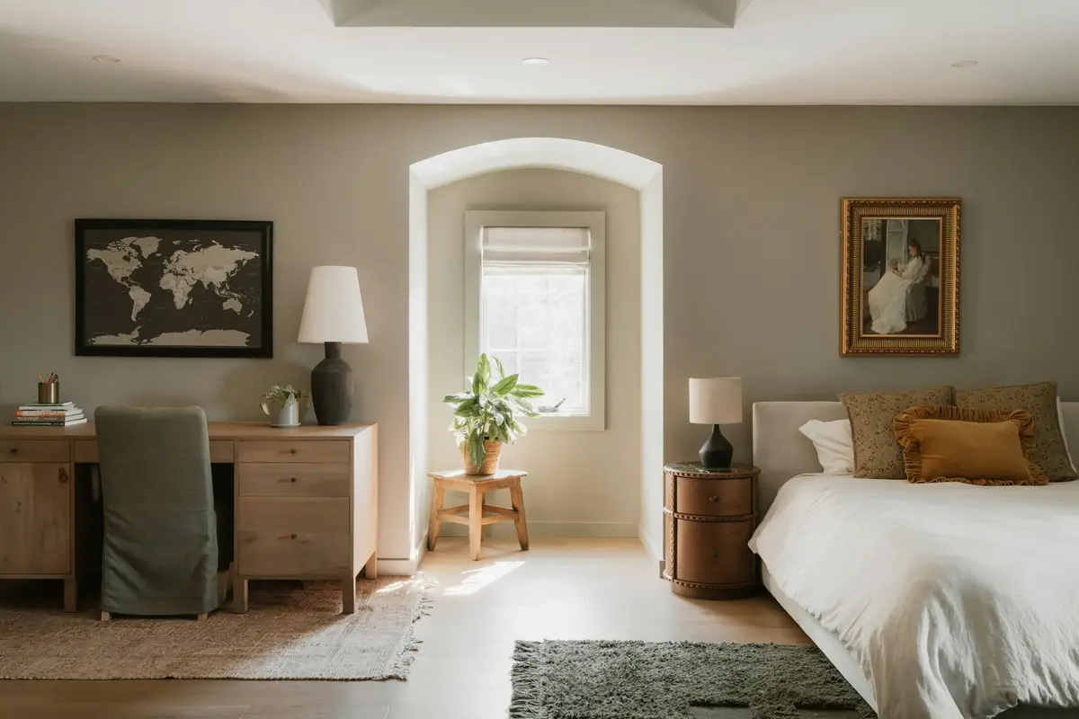Calm bedroom with a large bed in white linen, a wooden desk, an arched wall niche with a window and a plant, and framed pictures on beige walls